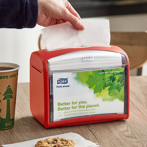 A person's hand using a red Tork Xpressnap tabletop napkin dispenser.