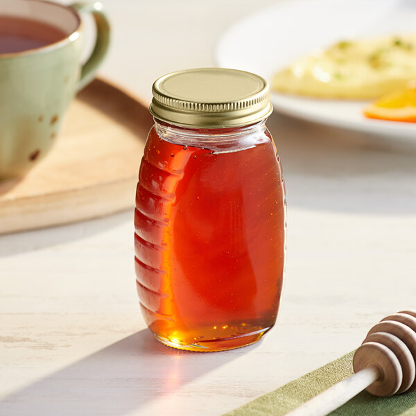 A Classic Queenline glass honey jar with a gold metal lid next to a cup of tea.