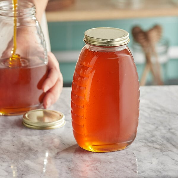 A person pouring honey into a Classic Queenline glass honey jar on a counter.