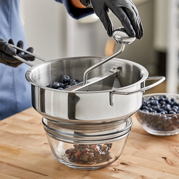 A person in gloves using a stainless steel food mill to strain blueberries.
