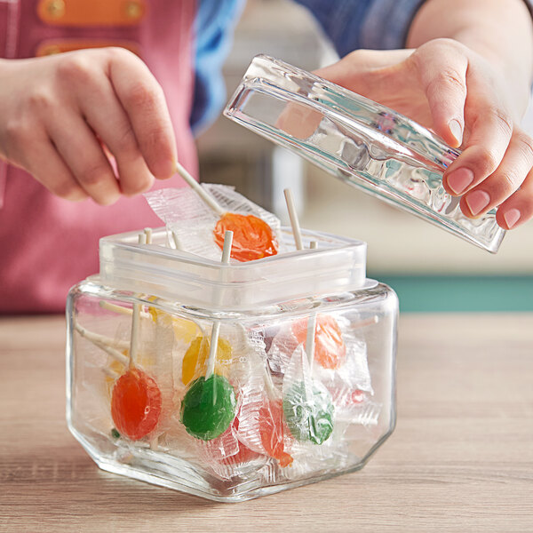 A person puts a lollipop into a clear Acopa glass jar on a kitchen counter.