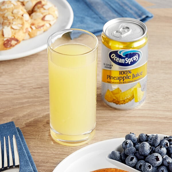 A glass of yellow liquid next to a can of Ocean Spray pineapple juice and a plate of food.