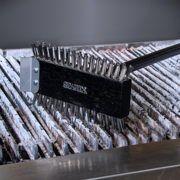 A Carlisle Sparta Spectrum metal grill brush with a scraper in use on a grill.