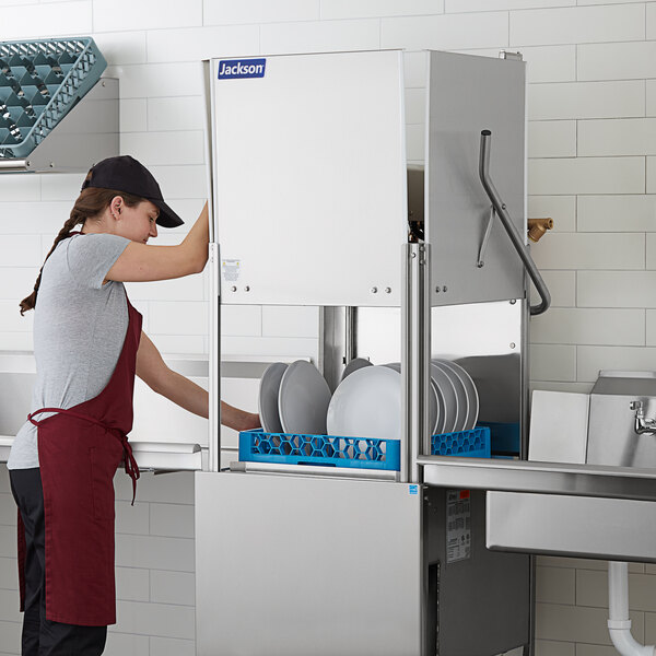 A woman in a red apron using a Jackson TempStar HH-E door type dishwasher to wash plates in a professional kitchen.