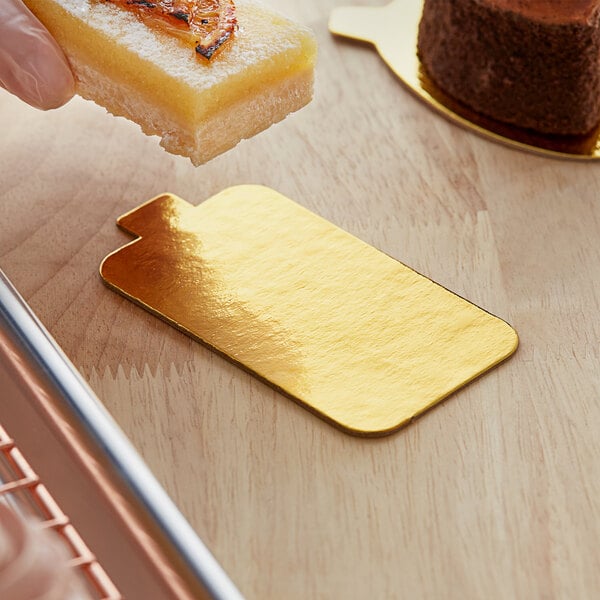 A hand holding a piece of food on a rectangular gold dessert board with a tab.