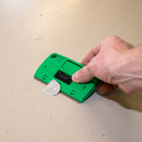 A hand using a Unger ProTrim 4" Glass Scraper with a green handle to clean a surface.