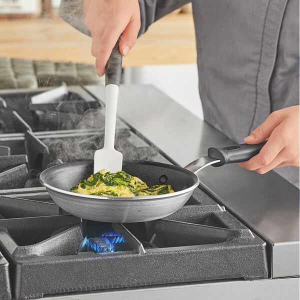 A person cooking food in a Vollrath Wear-Ever aluminum non-stick fry pan with a black silicone handle.