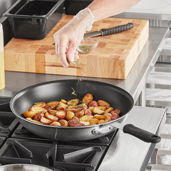 A person's hand in a black glove putting rosemary into a Vollrath stainless steel non-stick fry pan full of potatoes.