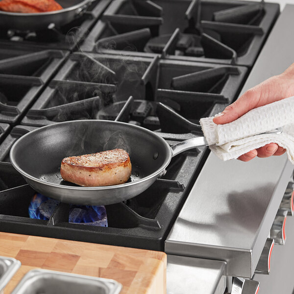 A person holding a Vollrath stainless steel frying pan with meat cooking in it.