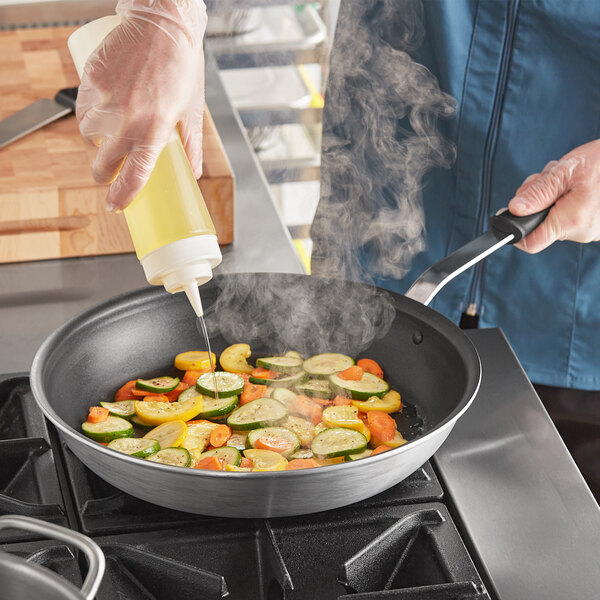 A person pouring yellow liquid into a Vollrath Wear-Ever non-stick frying pan filled with food.