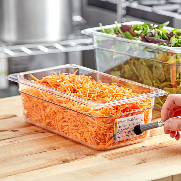 A hand uses a pen to label a clear plastic food pan of shredded carrots.