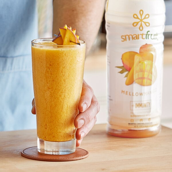 A hand holding a glass of yellow Smartfruit Mellow Mango beverage next to a bottle of Smartfruit on a table.