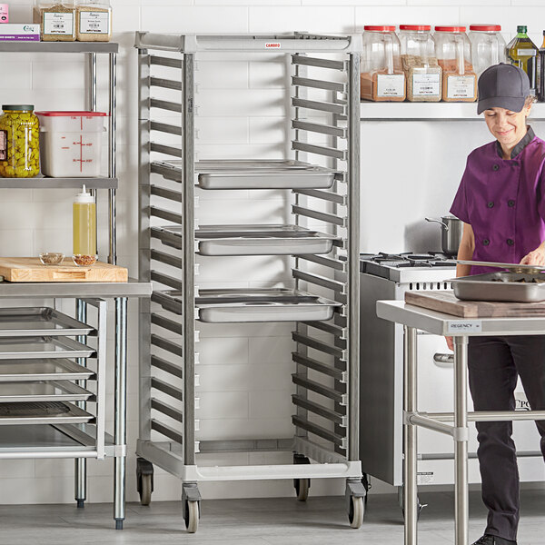 A woman in a kitchen rolling a metal tray rack filled with food.