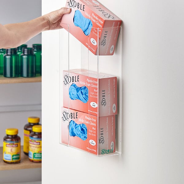 A hand using a Noble Products Triple Box Acrylic Wall-Mount Glove Dispenser to take a disposable glove from a box.