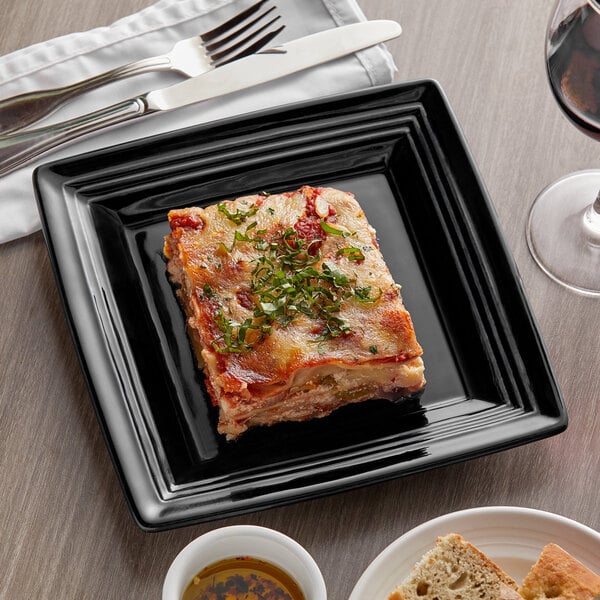 A Tuxton Concentrix black square china plate with a square piece of lasagna on it.