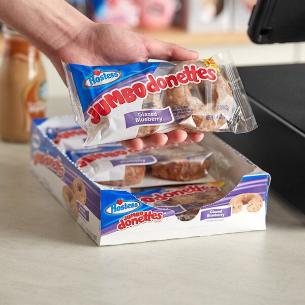 A hand holding a package of Hostess Glazed Blueberry Donettes in front of a cash register.