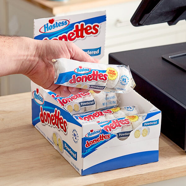 A hand holding a box of Hostess powdered donettes in front of a cash register.