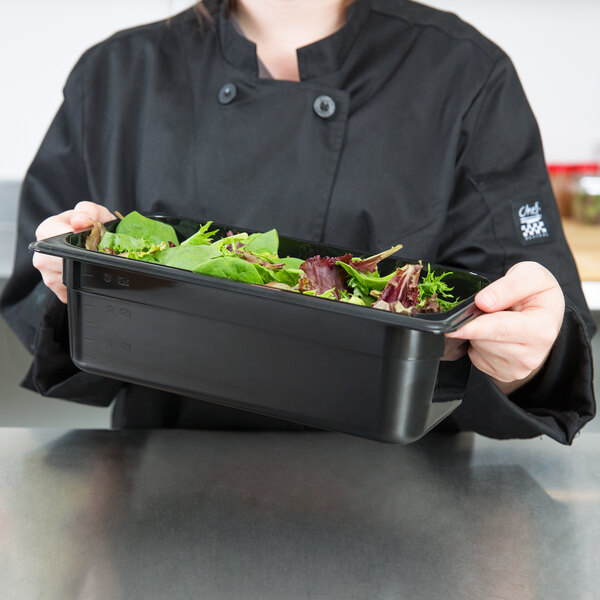 A woman in a chef's uniform holding a Cambro black polycarbonate food pan filled with salad.
