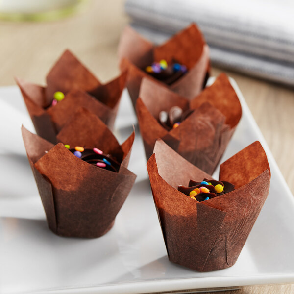 A group of brown Baker's Mark mini tulip baking cups on a white plate filled with chocolate cupcakes topped with colorful sprinkles.