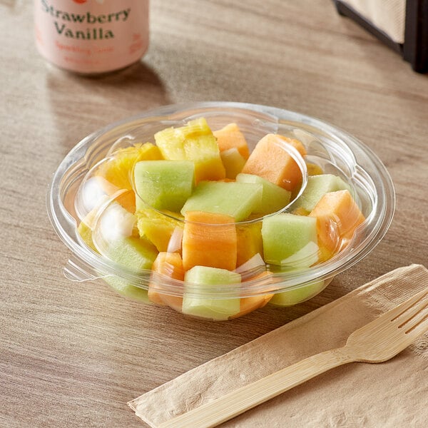 A clear plastic Choice salad bowl filled with sliced fruit with a wooden fork on the table.
