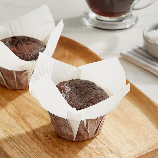 Two brown muffins wrapped in Baker's Mark white paper on a wooden tray.