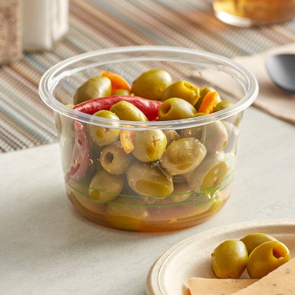 A Stalk Market clear plastic deli container filled with green olives and peppers on a counter with a plate of cheese.