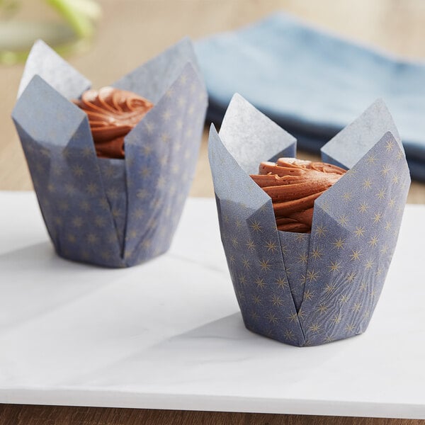Two Baker's Mark tulip baking cups holding cupcakes with blue and gold print wrappers on a table.