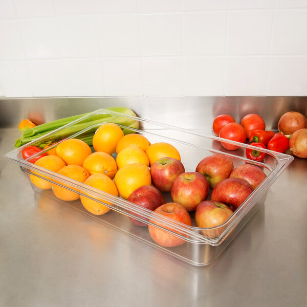 A Carlisle clear plastic food pan filled with oranges, apples, and celery on a counter.