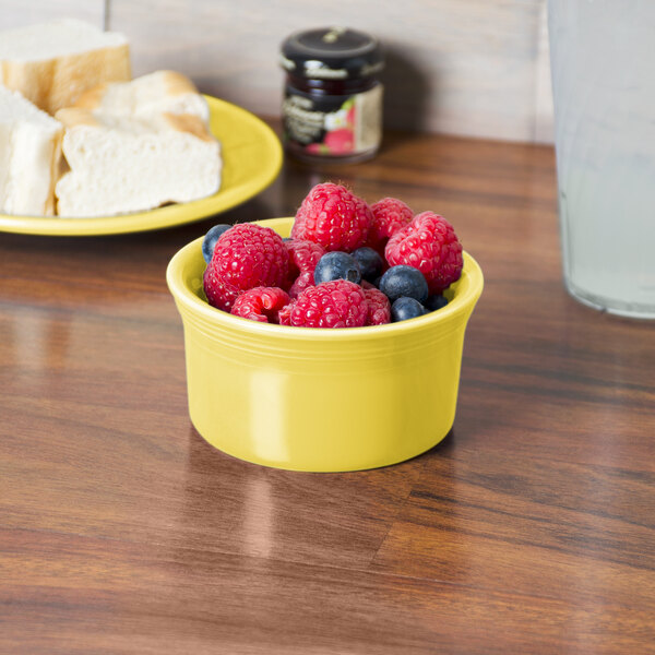 A yellow Fiesta ramekin filled with raspberries and blueberries on a white table.