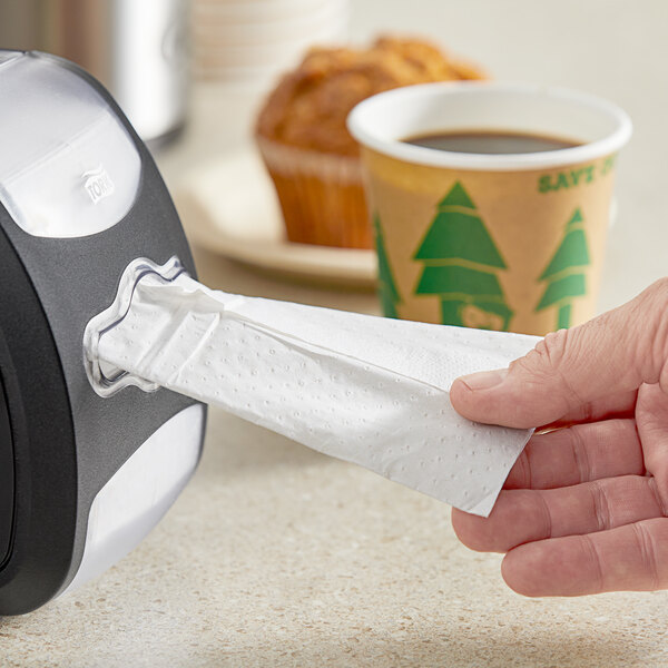 A hand pulling a Tork Xpressnap dispenser napkin from a dispenser on a counter next to a cup of coffee and a muffin.
