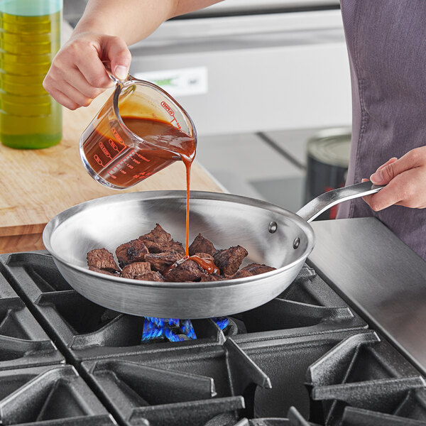 A person pouring sauce into a Vollrath Wear-Ever aluminum frying pan of meat.