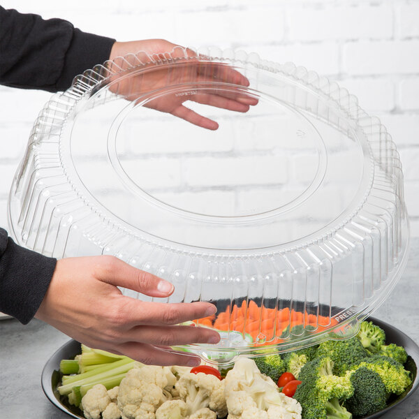 A person holding a WNA Comet clear plastic round high dome lid over a container of vegetables.