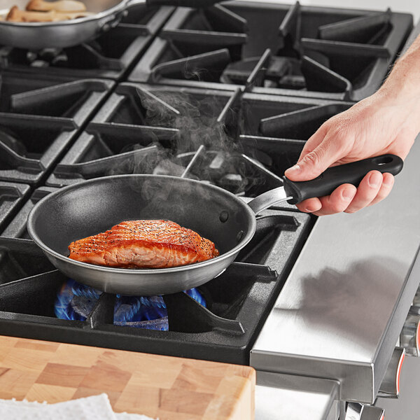 A person cooking a piece of salmon in a Vollrath stainless steel non-stick fry pan with a black silicone handle.