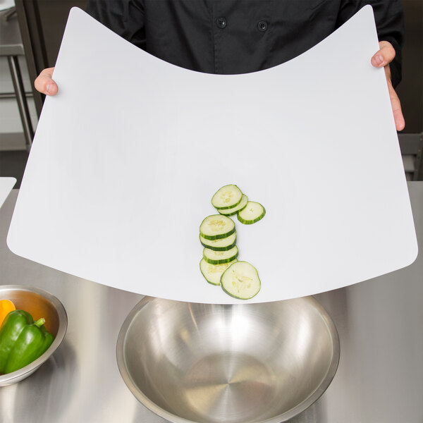 A chef using a Tablecraft white flexible cutting board to slice cucumbers on a white surface next to a bowl.