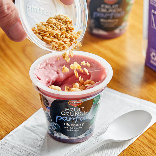 A hand pouring granola into a Del Monte Blueberry Parfait cup of yogurt.