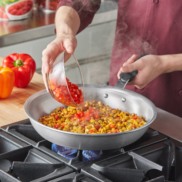 A person pouring a mixture of corn and red peppers into a Vollrath aluminum fry pan.