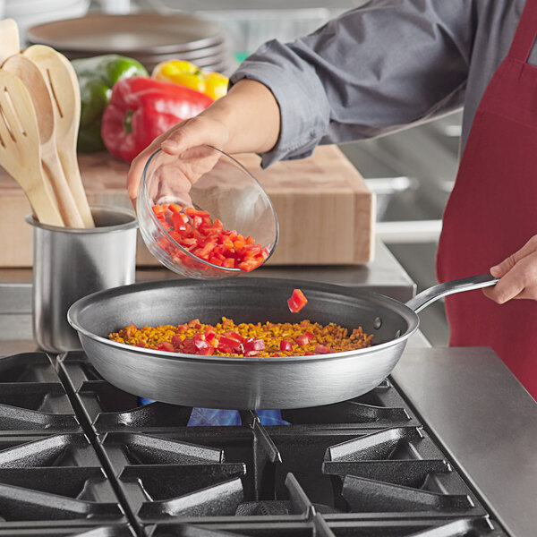 A woman pouring chopped red peppers into a Vollrath Wear-Ever non-stick fry pan on a stove.
