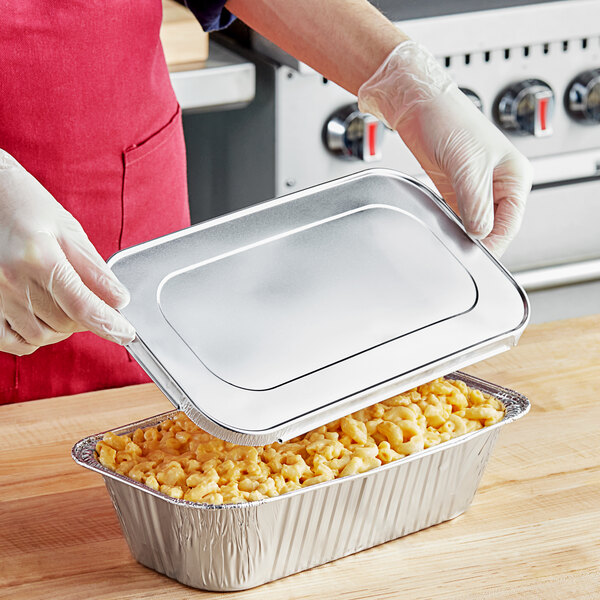 A person in gloves holding a Western Plastics foil steam table tray with macaroni and cheese with the lid open.