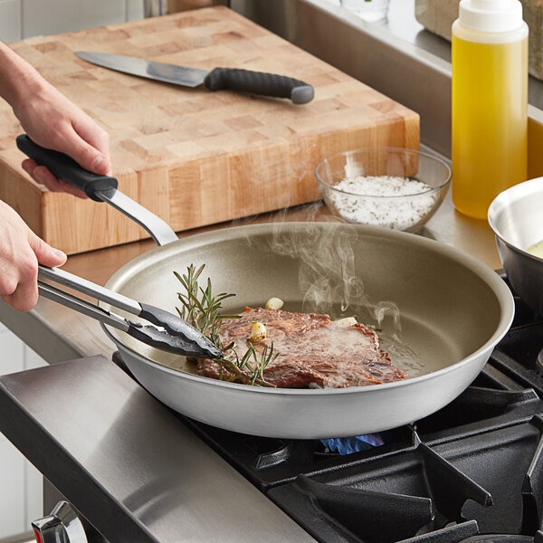 A piece of meat cooking in a Vollrath aluminum non-stick fry pan with a black silicone handle.