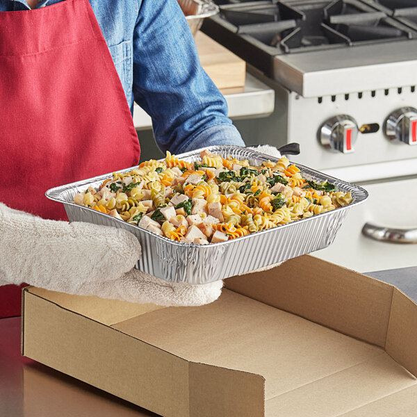 A person holding a Western Plastics foil steam table pan filled with pasta, chicken, and spinach.