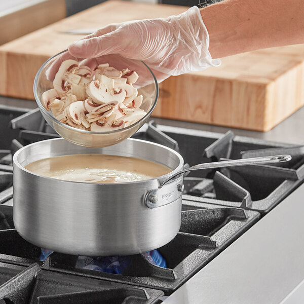 A person pouring a pot of mushroom soup into a Vollrath aluminum sauce pan on a stove.