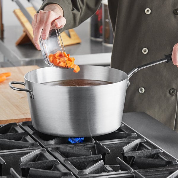 A person pouring carrots into a Vollrath Wear-Ever sauce pan on a gas stove.