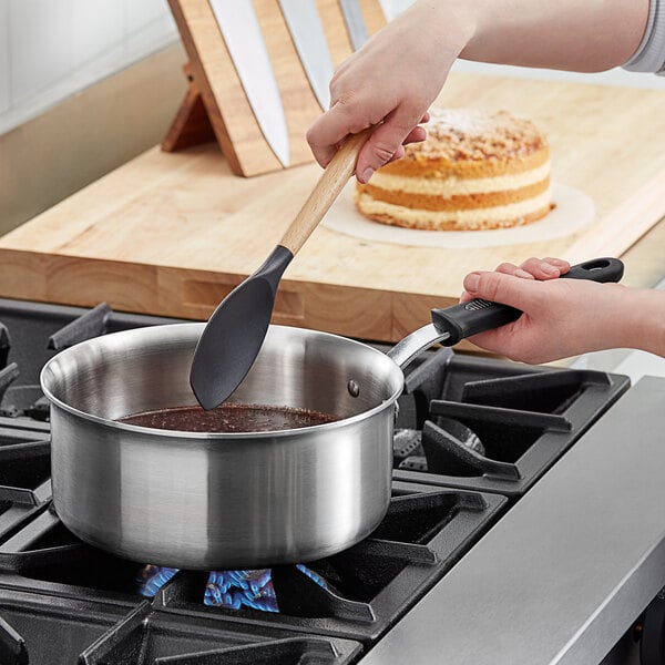 A person using a spatula to stir food in a Vollrath stainless steel sauce pan on a stove.