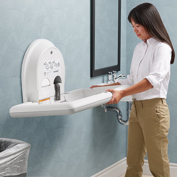 A woman holding a white rectangular Koala Kare baby changing station next to a sink.