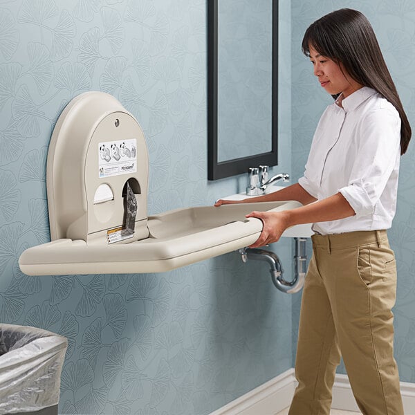 A woman standing next to a Koala Kare beige surface-mounted baby changing station.