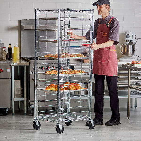 A man in a red apron standing next to a metal rack with trays of pastries.
