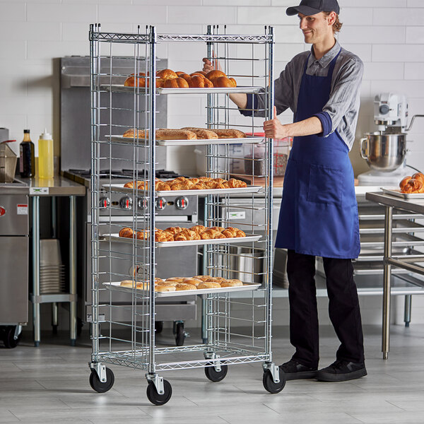 A man in a blue apron standing next to a Regency side load sheet pan rack filled with food.