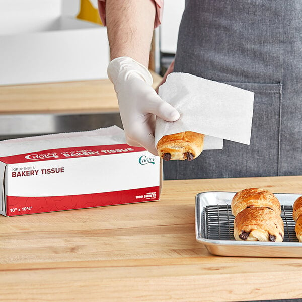 A person wearing white gloves using a pastry bag to decorate a pastry on a metal tray with a Choice bakery tissue.