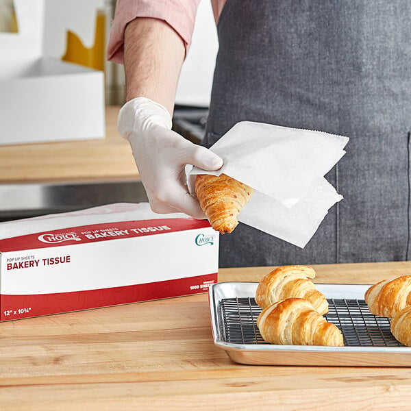 A hand holding a Choice bakery tissue sheet over a tray of croissants.