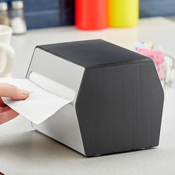 A person using a Dixie black and white tabletop napkin dispenser on a counter.